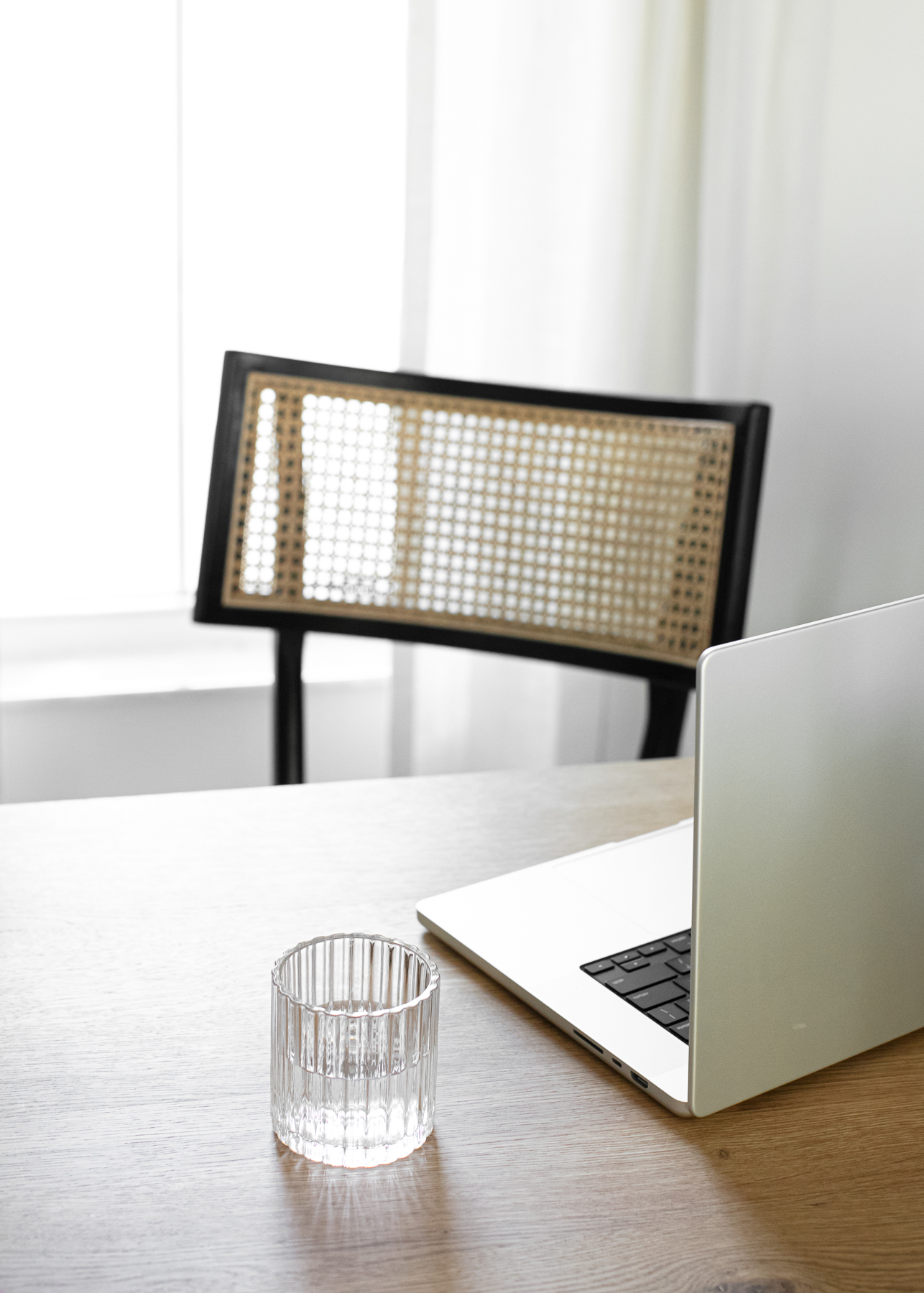 Minimal workspace with cane chair, glass of water, and laptop in soft morning light, creating a calm, focused environment for a midlife workday reset.